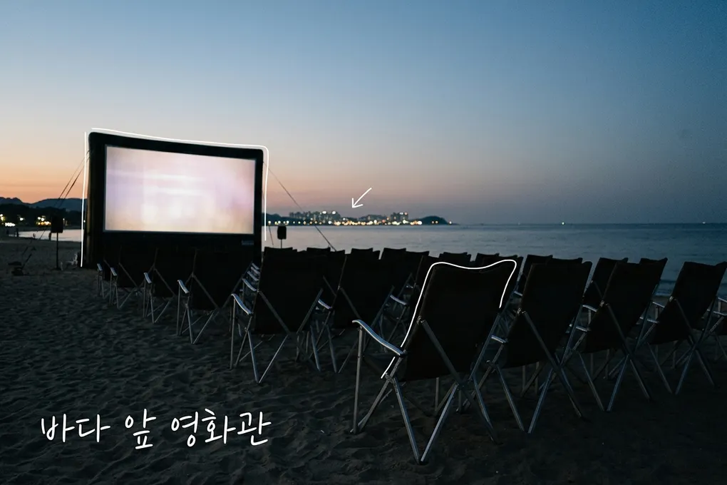 해 질 녘 바닷가 모래사장에 놓인 빈 접이식 의자들과 불이 켜진 야외 영화 스크린, 멀리 도시 불빛을 담은 AI 생성 이미지. 흰 선과 ‘바다 앞 영화관’ 손글씨가 얹혀 있다.
