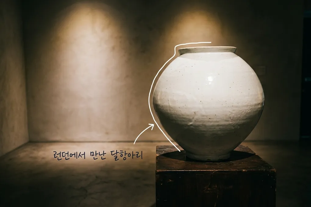 조명이 은은한 전시 공간의 낮은 받침 위에 놓인 흰 달항아리를 담은 AI 생성 이미지. 흰 선과 ‘런던에서 만난 달항아리’ 손글씨가 얹혀 있다. 실제 전시 사진이 아니다.