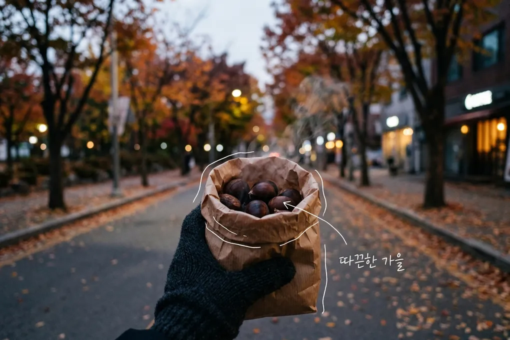 낙엽 쌓인 가로수길에서 장갑 낀 손이 김이 오르는 군밤 종이봉투를 든 AI 생성 이미지. 봉투에 흰 선이 얹혀 있고 '따끈한 가을' 손글씨가 있다.