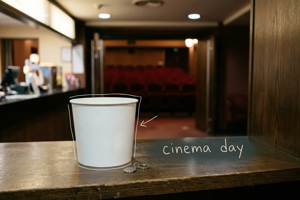 AI-generated image of a plain white popcorn bucket and a small keyring on a cinema lobby counter with blurred red seats behind. White pen lines trace the bucket and a handwritten note reads 'cinema day'.
