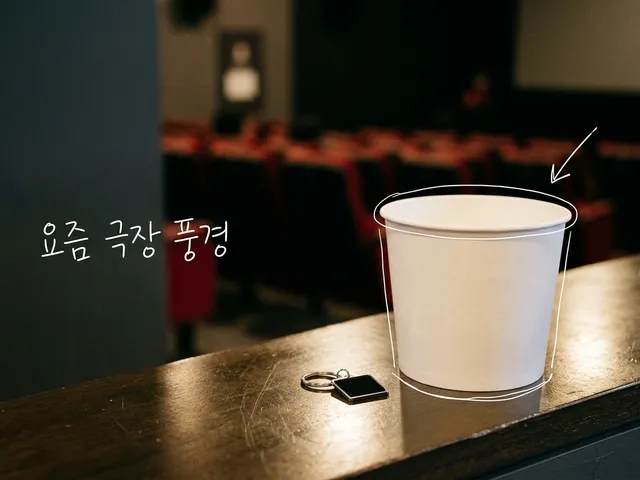 극장 로비 카운터 위에 놓인 흰 팝콘통과 작은 키링, 뒤로 흐릿한 객석이 보이는 AI 생성 이미지. 팝콘통에 흰 선이 얹혀 있고 '요즘 극장 풍경' 손글씨가 있다.