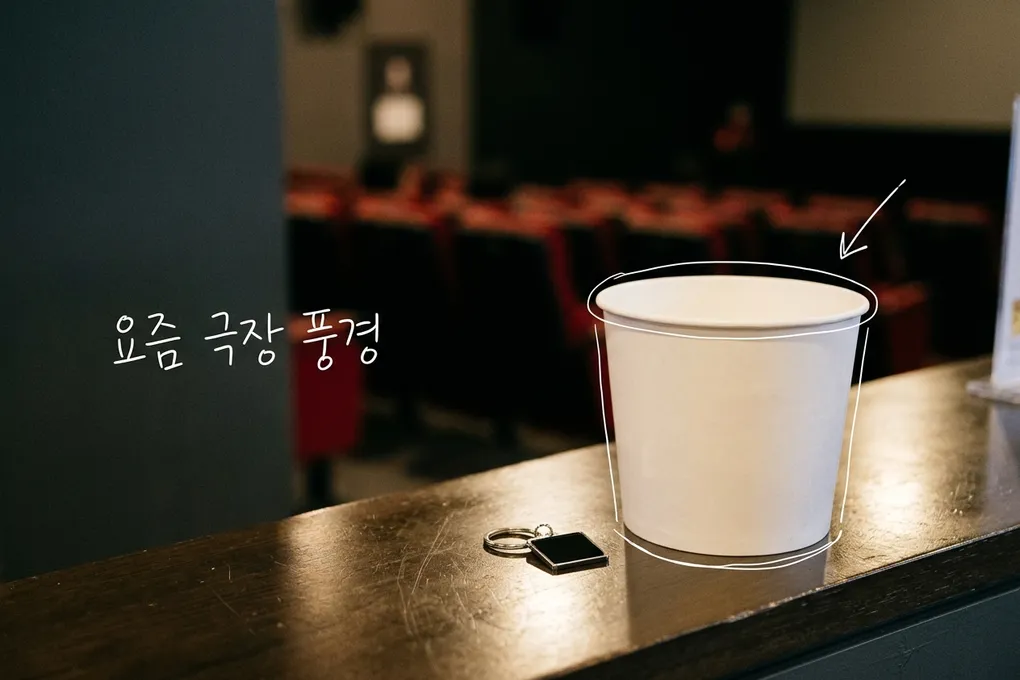 극장 로비 카운터 위에 놓인 흰 팝콘통과 작은 키링, 뒤로 흐릿한 객석이 보이는 AI 생성 이미지. 팝콘통에 흰 선이 얹혀 있고 '요즘 극장 풍경' 손글씨가 있다.