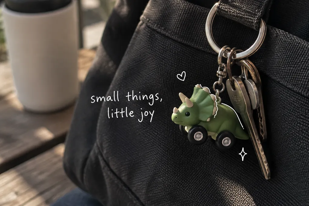 AI-generated green dinosaur car charm on a dark canvas bag, with a small white heart and the handwritten words small things, little joy.