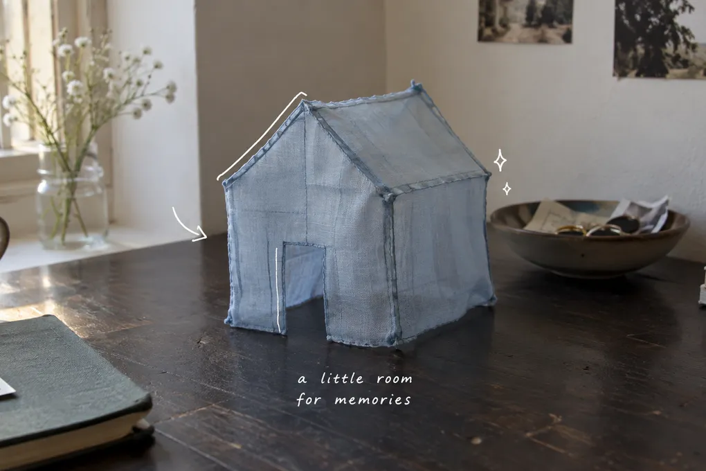 AI-generated still life of a small blue cloth house on a wooden table, with thin white lines and a note about memories.