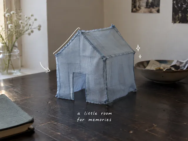 AI-generated still life of a small blue cloth house on a wooden table, with thin white lines and a note about memories.