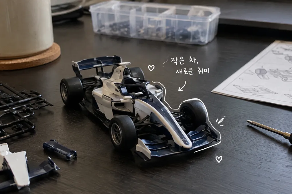 책상 위 모형 경주차와 작은 조립 도구에 흰 손글씨를 더한 AI 생성 이미지.