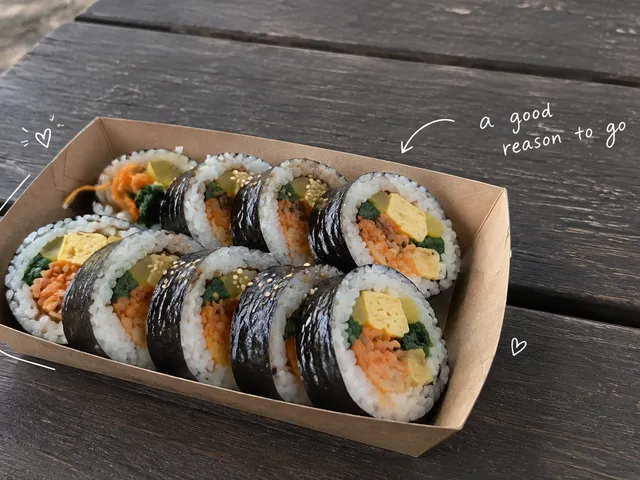 AI-generated sliced gimbap in a paper tray on a wooden picnic table, with a short white handwritten note.