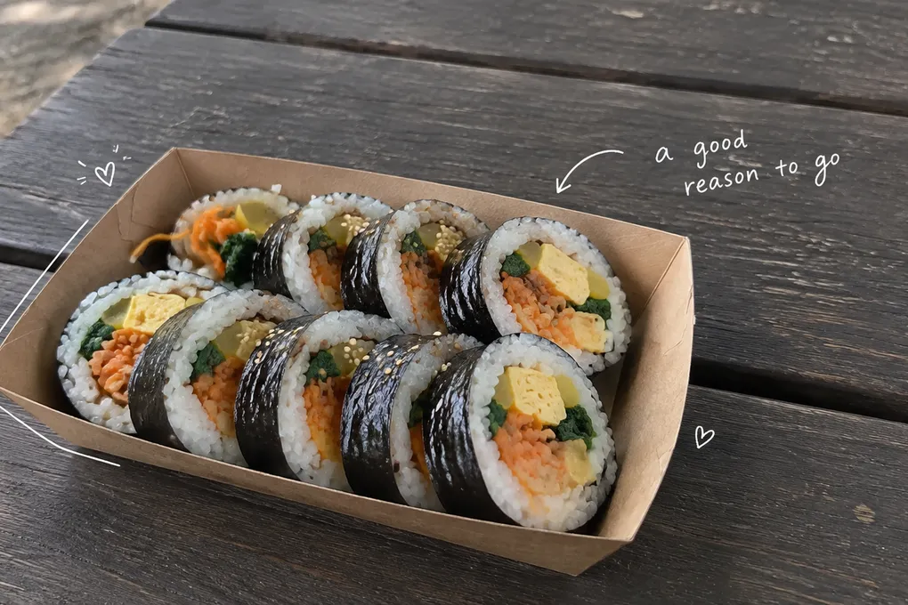 AI-generated sliced gimbap in a paper tray on a wooden picnic table, with a short white handwritten note.