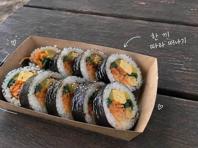 종이 트레이에 담긴 썬 김밥이 나무 피크닉 테이블에 놓인 AI 생성 이미지와 짧은 흰 손글씨.
