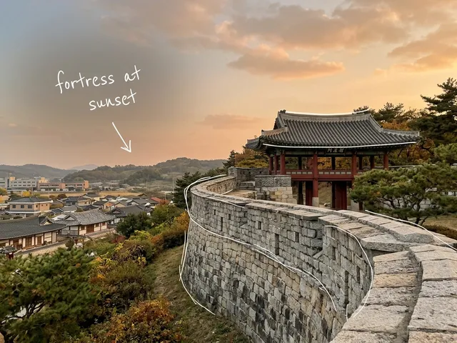 AI-generated sunset view of a stone fortress wall and tiled-roof pavilion above a Korean town, with thin white lines and the handwritten note fortress at sunset.