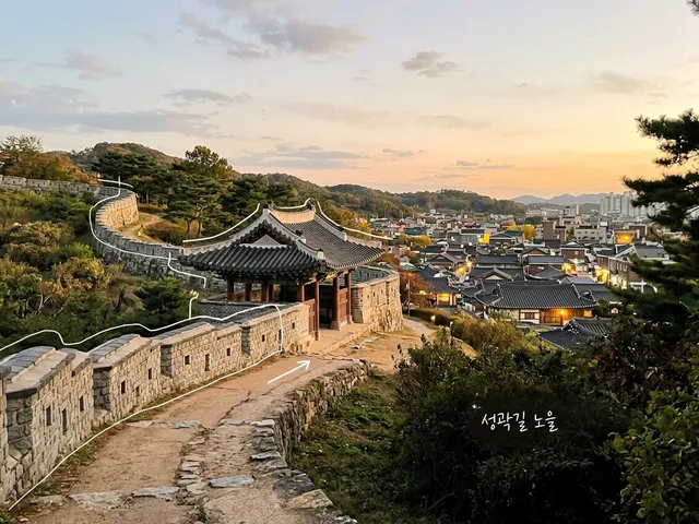 해 질 녘 돌성곽과 기와 문루, 아래로 기와지붕 마을이 보이는 AI 생성 이미지. 흰 선과 ‘성곽길 노을’ 손글씨가 얹혀 있다.