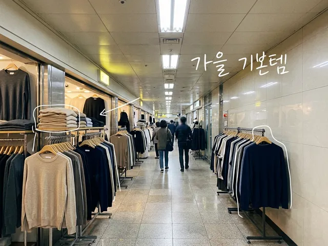 지하상가 통로 양쪽에 베이지·회색·남색 니트와 티셔츠가 걸린 AI 생성 이미지. 쌓인 니트와 옷걸이에 흰 선이 얹혀 있고 '가을 기본템' 손글씨가 있다.