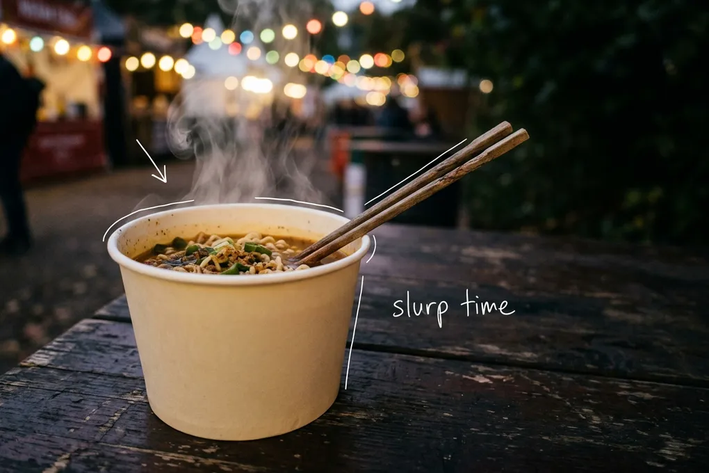 AI-generated steaming paper cup of ramyeon with wooden chopsticks on an outdoor festival table, with blurred festival lights behind. White lines trace the cup rim and steam, with the handwritten note slurp time.