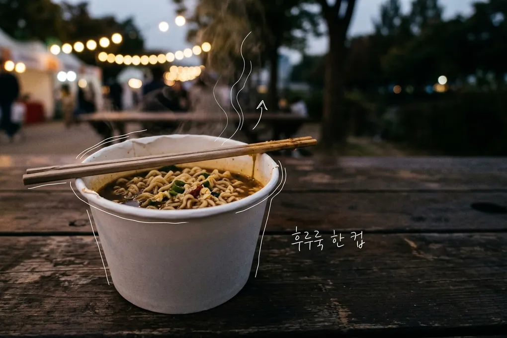 야외 축제장 나무 테이블 위에서 김이 오르는 종이컵 라면과 나무젓가락, 뒤로 흐릿한 축제 조명을 담은 AI 생성 이미지. 컵 테두리와 김에 흰 선이 얹혀 있고 '후루룩 한 컵' 손글씨가 있다.