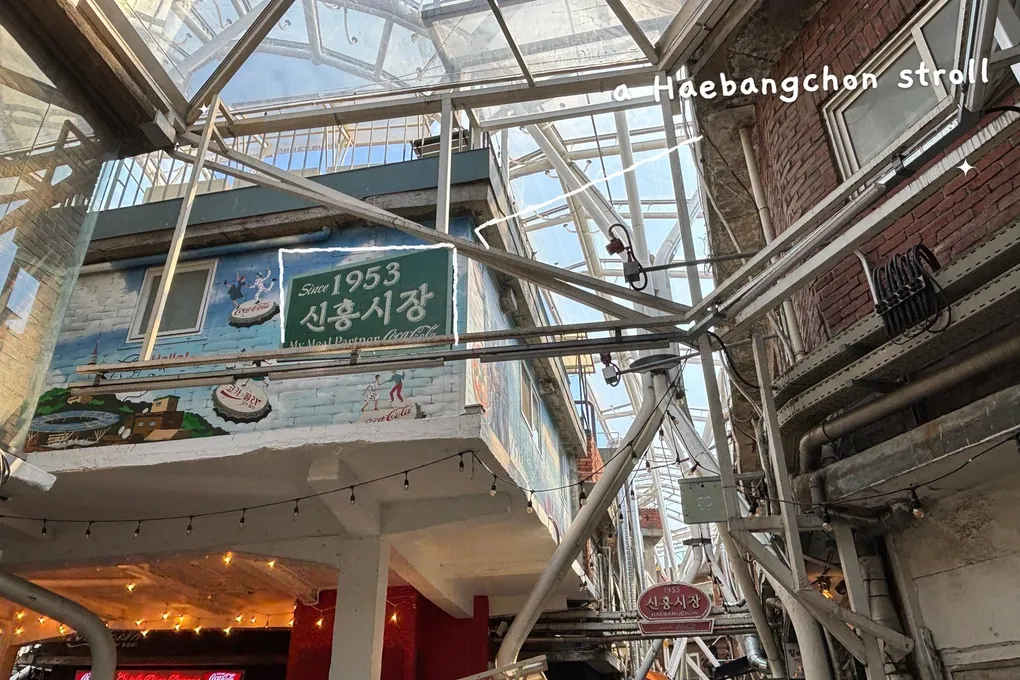 A real photo of the 'Since 1953 Sinheung Market' mural sign under the clear roof of the market alley, with thin white hand-drawn lines around the sign and the handwritten note 'a Haebangchon stroll'.