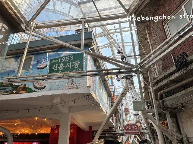 A real photo of the 'Since 1953 Sinheung Market' mural sign under the clear roof of the market alley, with thin white hand-drawn lines around the sign and the handwritten note 'a Haebangchon stroll'.