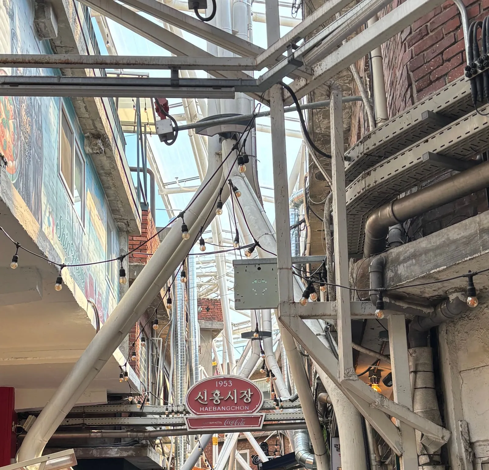 The 'Since 1953 Sinheung Market Haebangchon' sign at the market entrance, with tangled pipes and sky seen through the clear roof