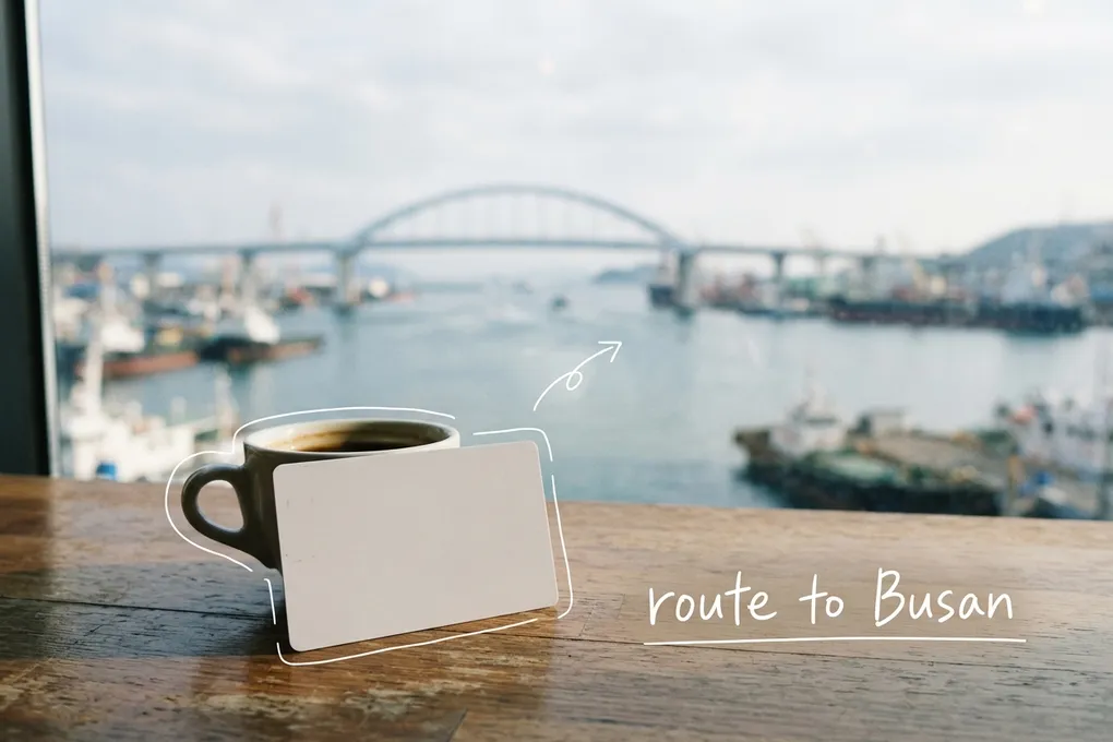 AI-generated blank white transit card standing beside a cup on a cafe table, with a harbor bridge outside the window, thin white lines and the handwritten note 'route to Busan'.