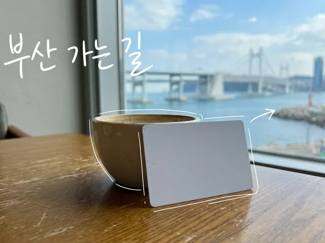 창가 카페 테이블 위 컵 옆에 세워진 흰색 빈 교통카드와 창밖의 항구 다리를 담은 AI 생성 이미지. 흰 선과 ‘부산 가는 길’ 손글씨가 얹혀 있다.