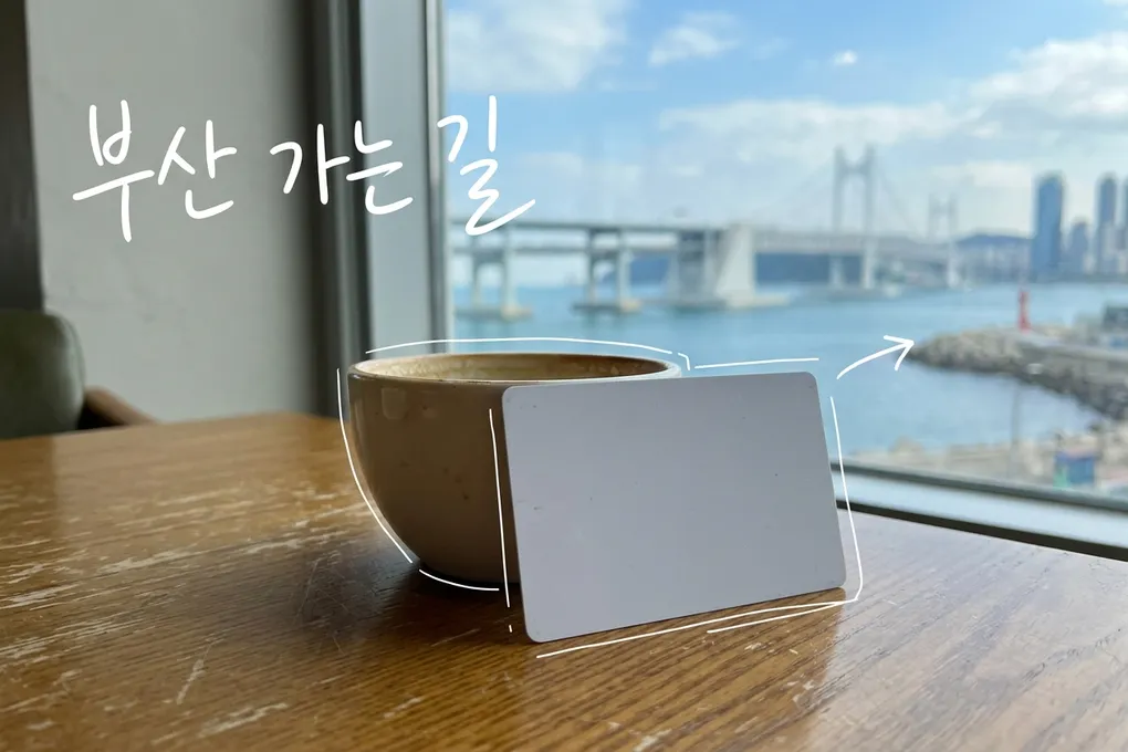 창가 카페 테이블 위 컵 옆에 세워진 흰색 빈 교통카드와 창밖의 항구 다리를 담은 AI 생성 이미지. 흰 선과 ‘부산 가는 길’ 손글씨가 얹혀 있다.