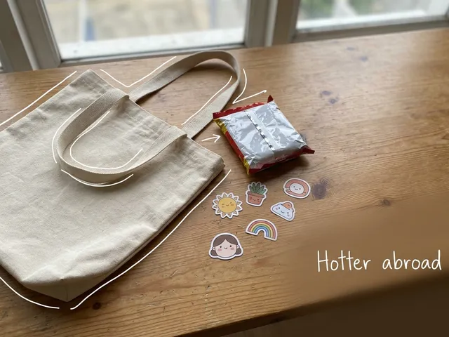 AI-generated image of a canvas tote bag, an instant noodle packet and cute stickers on a wooden table. A thin white line traces the bag next to a handwritten note reading 'Hotter abroad'.