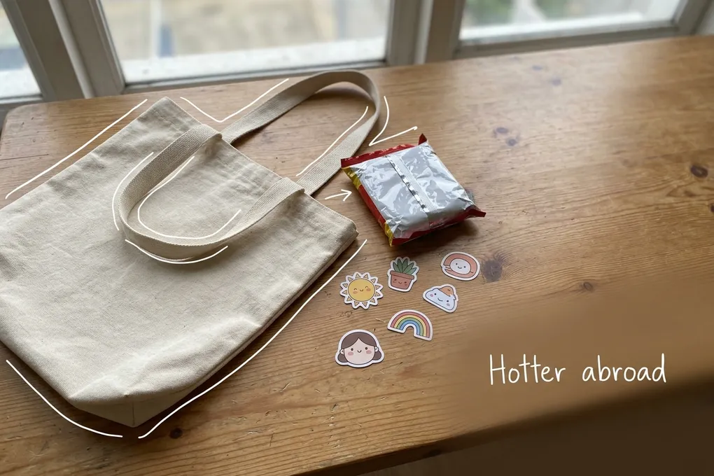 AI-generated image of a canvas tote bag, an instant noodle packet and cute stickers on a wooden table. A thin white line traces the bag next to a handwritten note reading 'Hotter abroad'.