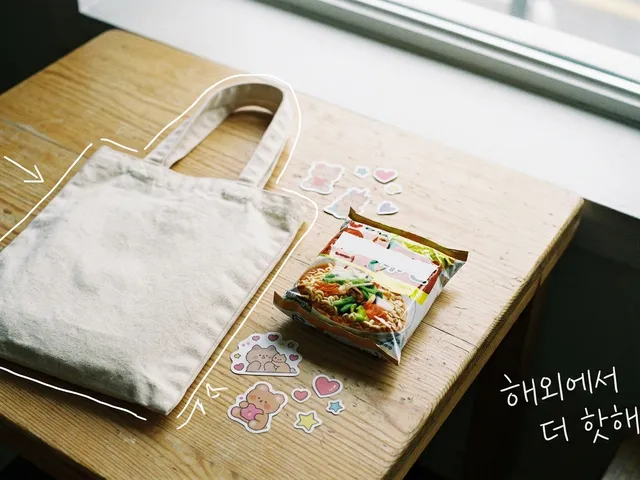 나무 탁자 위에 캔버스 장바구니, 라면 봉지, 귀여운 스티커가 놓인 AI 생성 이미지. 장바구니에 흰 선과 '해외에서 더 핫해' 손글씨가 얹혀 있다.