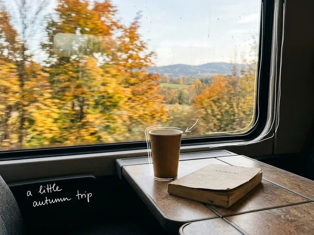 AI-generated view from a train window seat: a paper cup and a book on the fold-down table, autumn leaves rushing past outside, with thin white lines and the handwritten note 'a little autumn trip'.