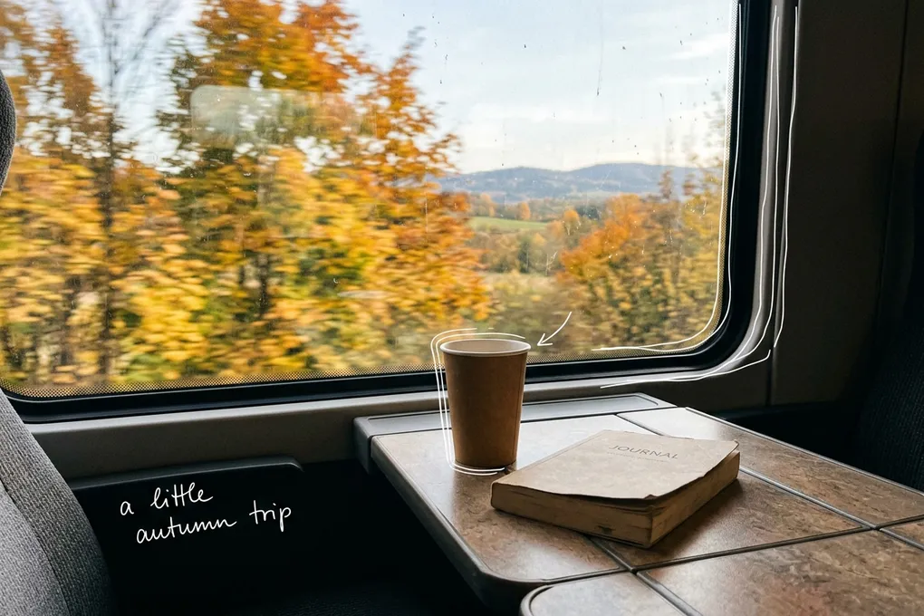 AI-generated view from a train window seat: a paper cup and a book on the fold-down table, autumn leaves rushing past outside, with thin white lines and the handwritten note 'a little autumn trip'.