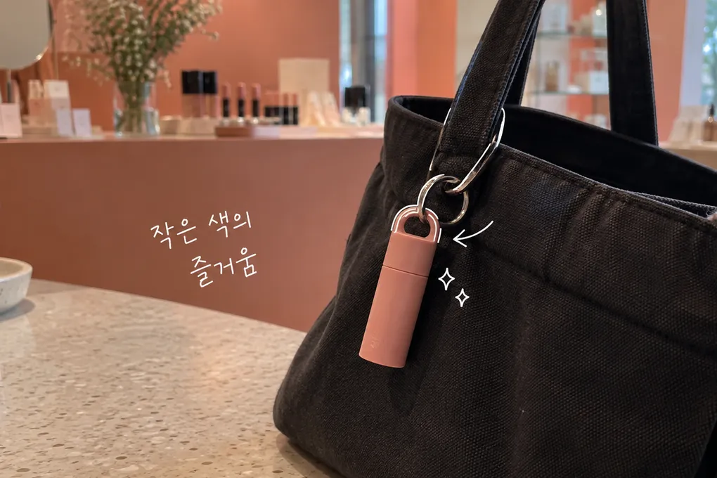 검은 가방에 매달린 코랄색 립밤과 흰 펜 선, 조금의 색이라는 손글씨를 담은 AI 생성 일러스트.