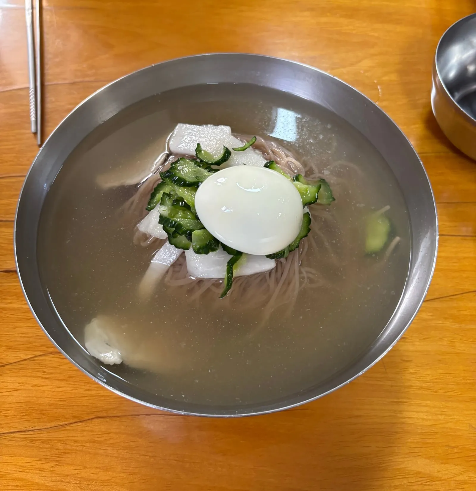 Cold noodles in a stainless steel bowl, topped with cucumber, pale vegetable slices and half a boiled egg