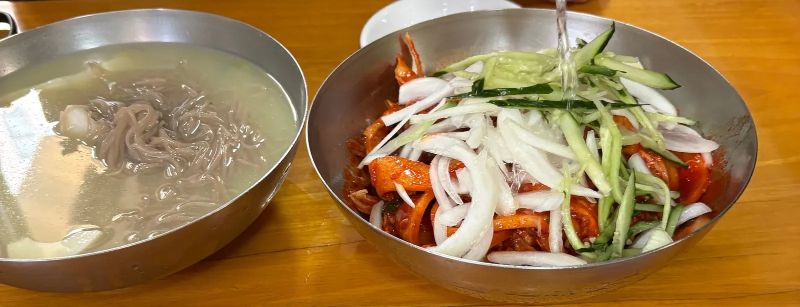 A table with cold noodles and spicy cold noodles, with broth being poured over the spicy bowl