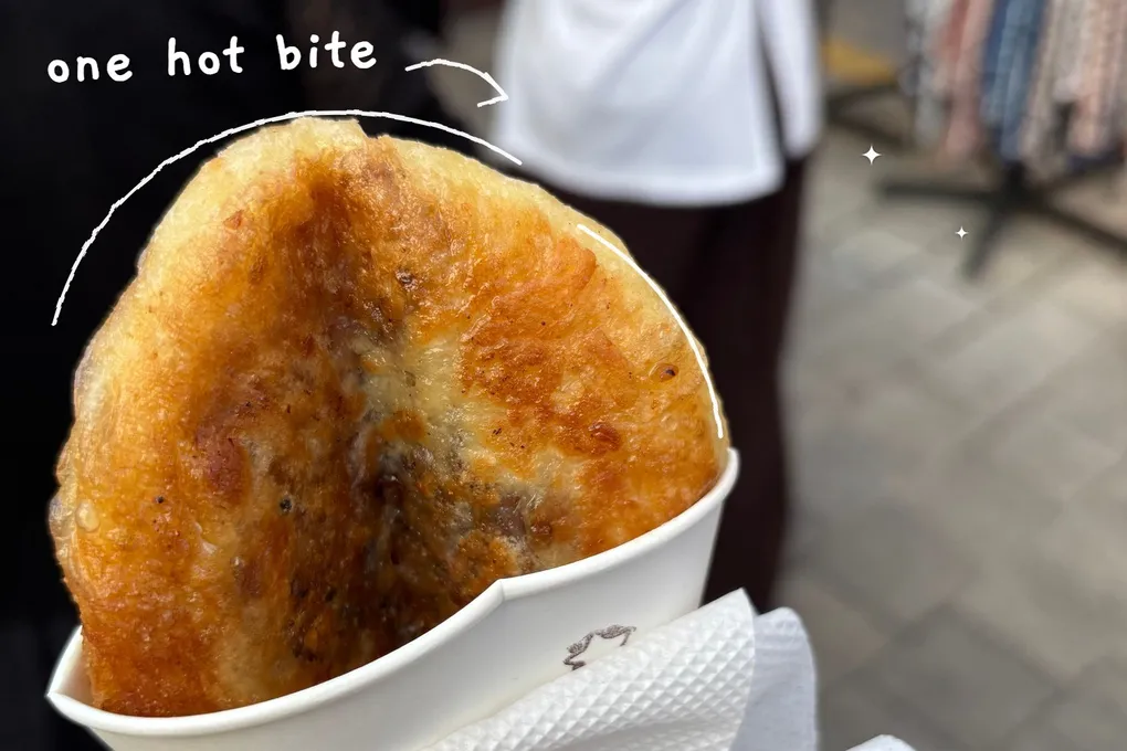 A real photo of a golden fried hotteok in a paper cup, with thin white hand-drawn lines following its outline and the handwritten note 'one hot bite'.