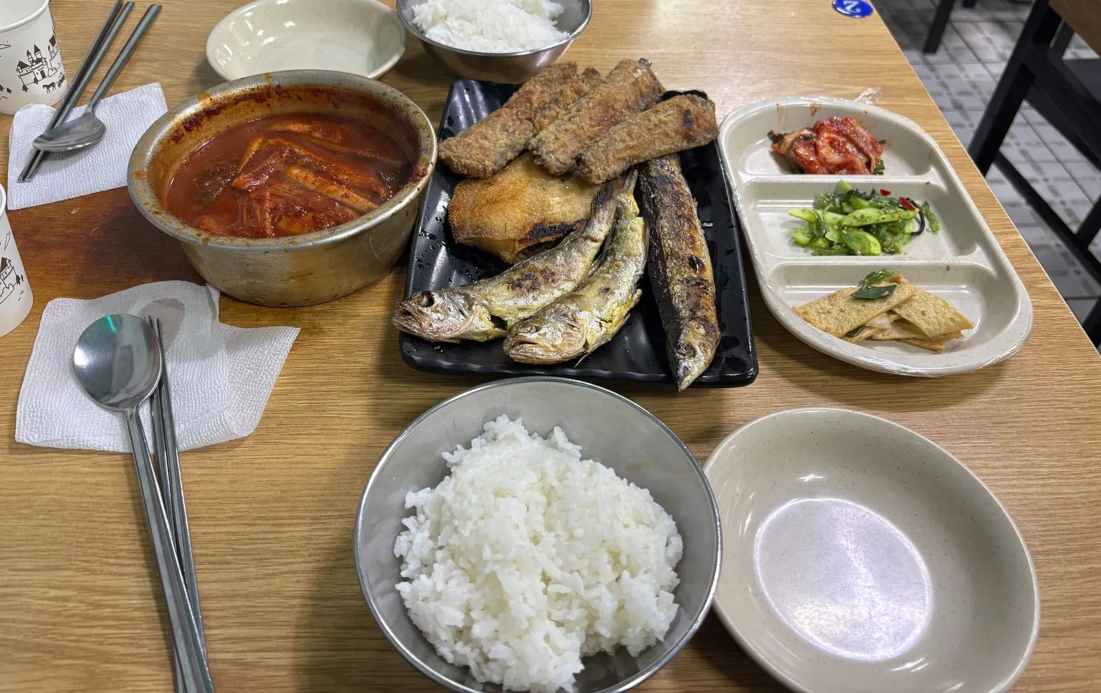 A restaurant table with braised hairtail in an aluminum pot, grilled fish on a black plate, rice and side dishes