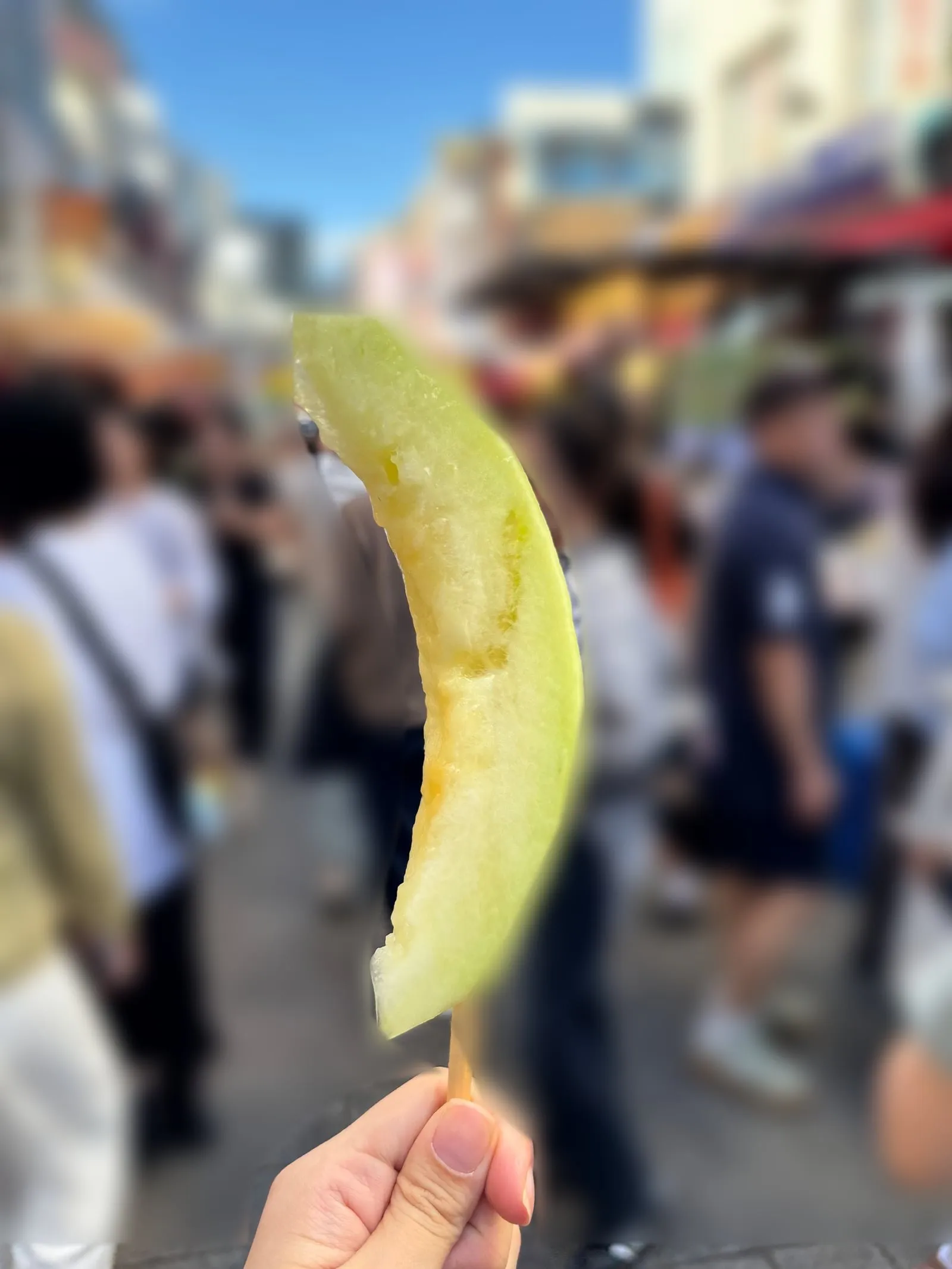 A hand holding a melon skewer, with the people in the background blurred