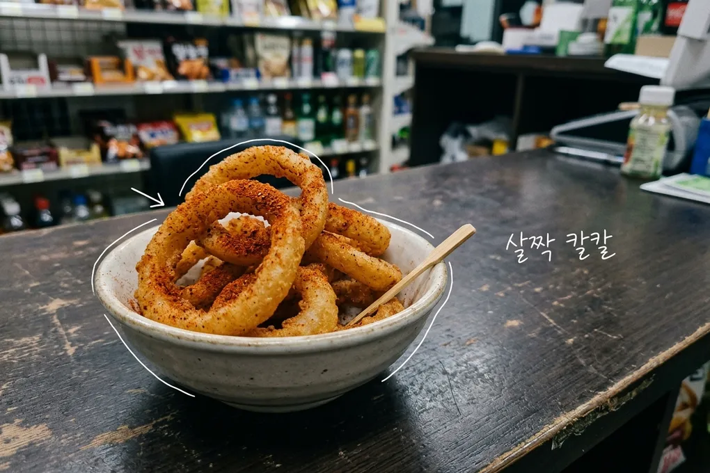 나무 카운터 위 그릇에 붉은 양념 가루를 뿌린 양파링 모양 과자가 담긴 AI 생성 이미지. 흰 선과 ‘살짝 칼칼’ 손글씨가 얹혀 있다.