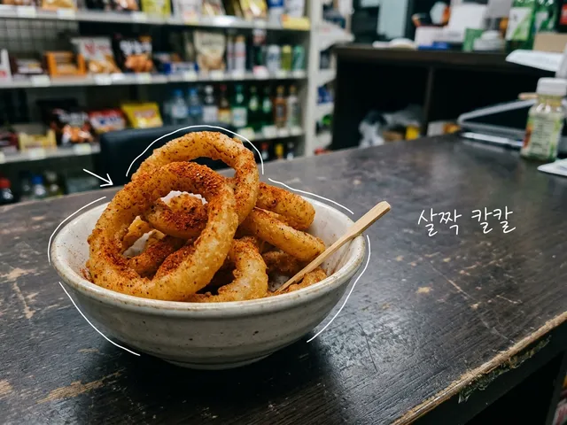 나무 카운터 위 그릇에 붉은 양념 가루를 뿌린 양파링 모양 과자가 담긴 AI 생성 이미지. 흰 선과 ‘살짝 칼칼’ 손글씨가 얹혀 있다.