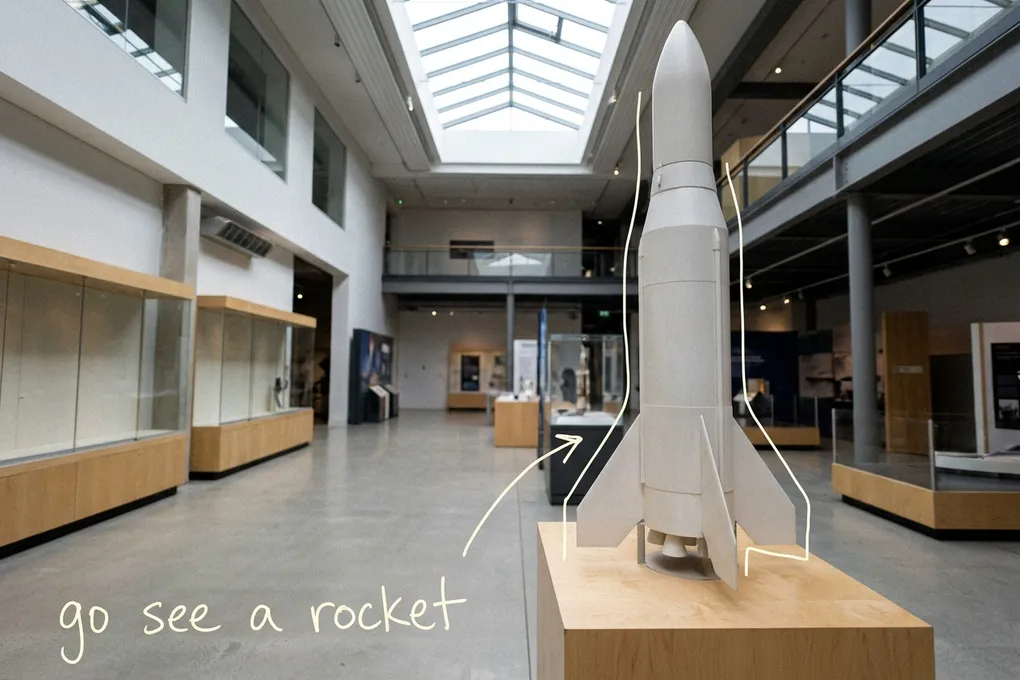 AI-generated white scale-model rocket on a wooden stand in a bright museum hall with a skylight, with thin white lines and the handwritten note go see a rocket; not a photo of the real museum.