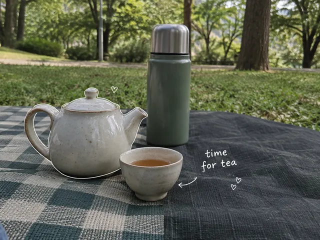 AI-generated small teapot, filled cup and thermos on a park picnic mat, with a white time-for-tea note.