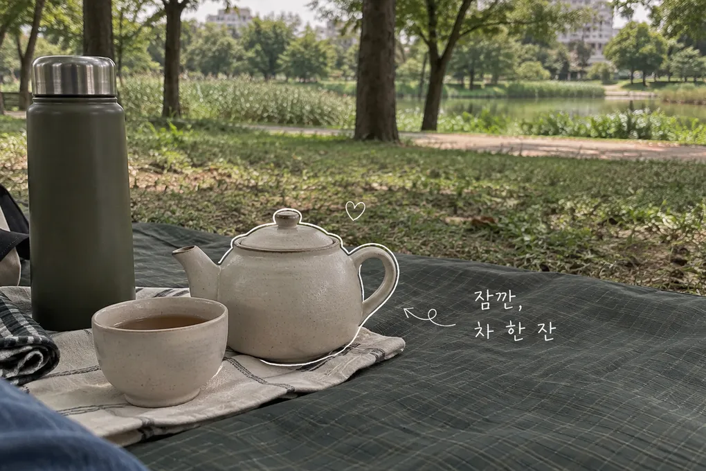 공원 돗자리 위 작은 다관과 찻잔, 보온병에 흰 손글씨를 더한 AI 생성 이미지.