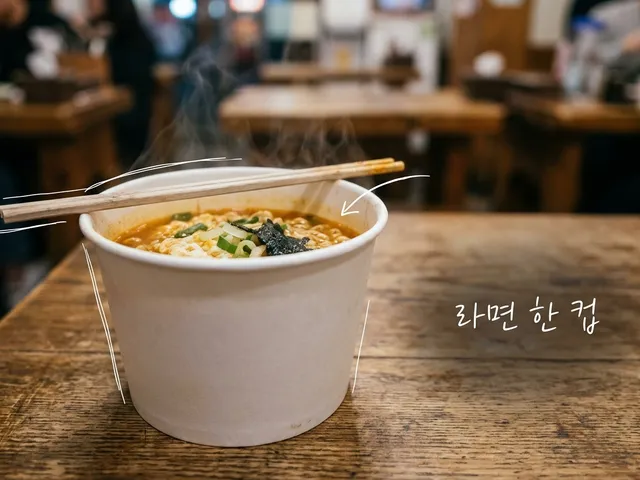 나무 테이블 위에 젓가락을 걸친 무지 종이컵 라면에서 김이 오르는 모습을 담은 AI 생성 이미지. 흰 선과 ‘라면 한 컵’ 손글씨가 얹혀 있다.