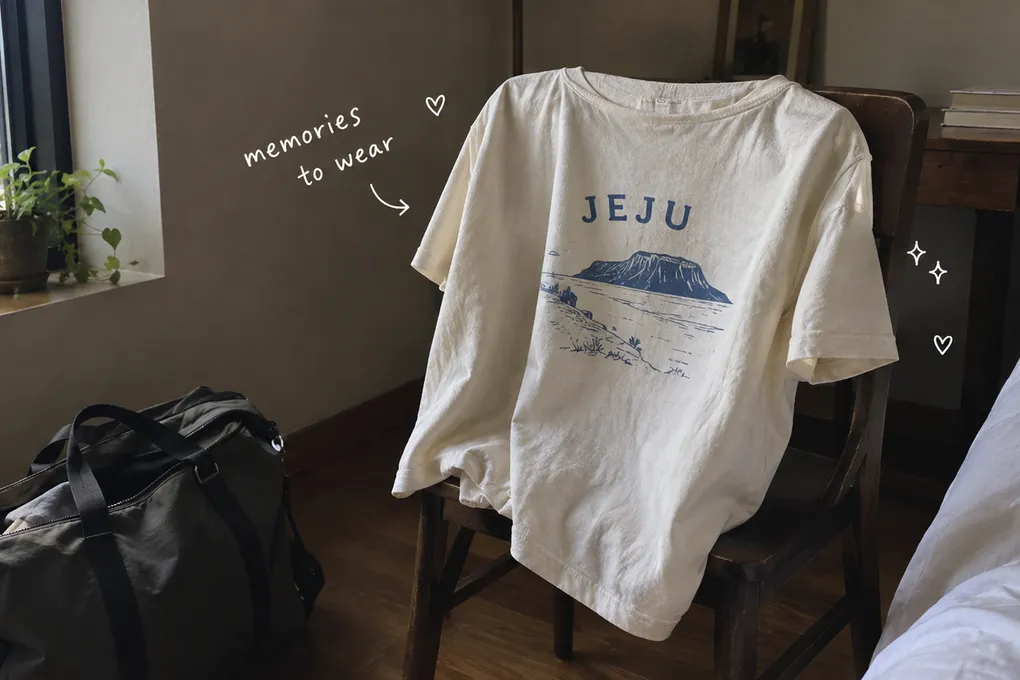 AI-generated cream Jeju souvenir T-shirt draped over a wooden chair beside a travel bag, with handwritten words memories to wear.