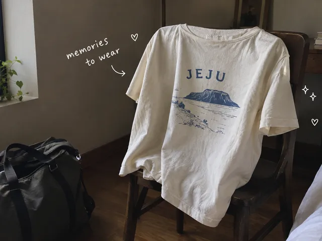AI-generated cream Jeju souvenir T-shirt draped over a wooden chair beside a travel bag, with handwritten words memories to wear.