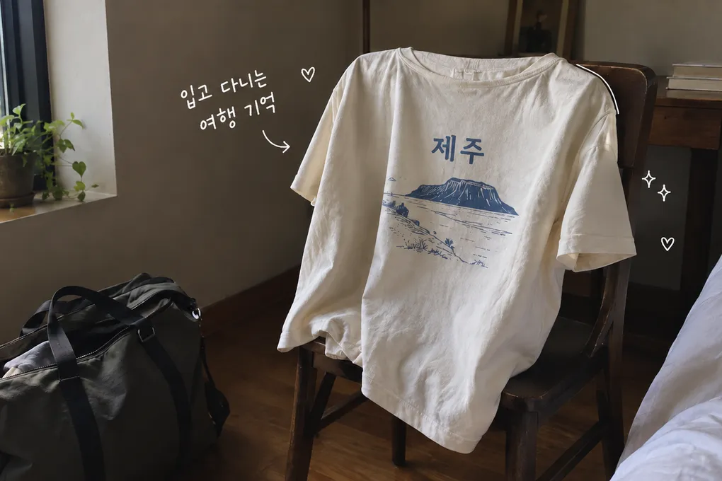 나무 의자에 걸친 제주 기념 티셔츠와 여행 가방, 입고 다니는 여행 기억이라는 손글씨를 담은 AI 생성 이미지.